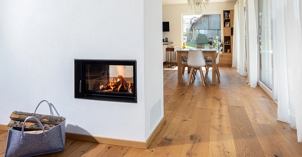 Kaminofen im Einsatz in einem modernen Wohnzimmer mit weiß gestrichenen Wänden und Holzboden. Ein Korb mit Brennholz steht daneben. Im Hintergrund ein Esstisch mit Stühlen und großen Fenstern.