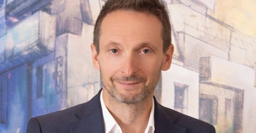 Großaufnahme eines mitteljungen Mannes mit kurzem braunen haar und im Anzug ohne Krawatte, er steht vor einem Bild mit Großstadtansicht.