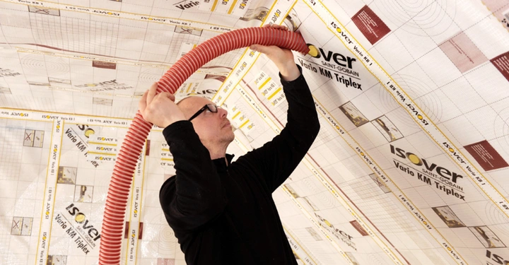 Eine Person installiert einen roten Wellschlauch unter einer mit Dämmfolie von Isover bespannten Dachschräge. Text auf Folie: Isover Saint-Gobain Vario KM Triplex.