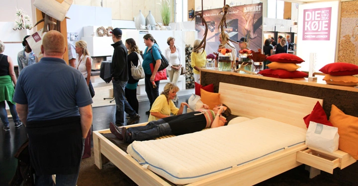 Personen testen ein Bett in einer Ausstellungsumgebung es gibt eine Anzeige mit dem Text DIE KOJE BETT UND ZUG CHRISTIAN LEGMINGER und mehrere Besucher betrachten die Ausstellung objekten