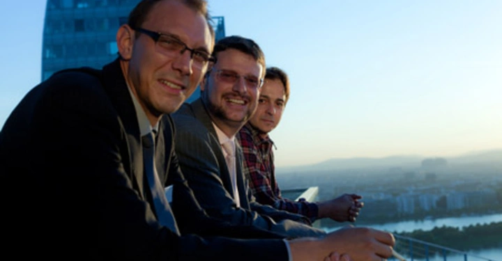 Galerie-Bild 3: Drei Männer lehnen auf einem Balkon und lächeln in die Kamera Hochhaus im Hintergrund Stadtlandschaft mit Fluss im Sonnenuntergang.
