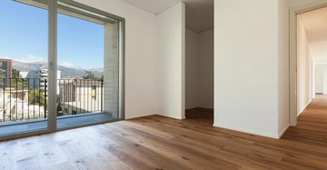Neuer Raum mit Holzboden Action ohne Einrichtung umgeben von weißen Wänden großem Fenster und Balkonblick auf Gebäude und Berge im Hintergrund.