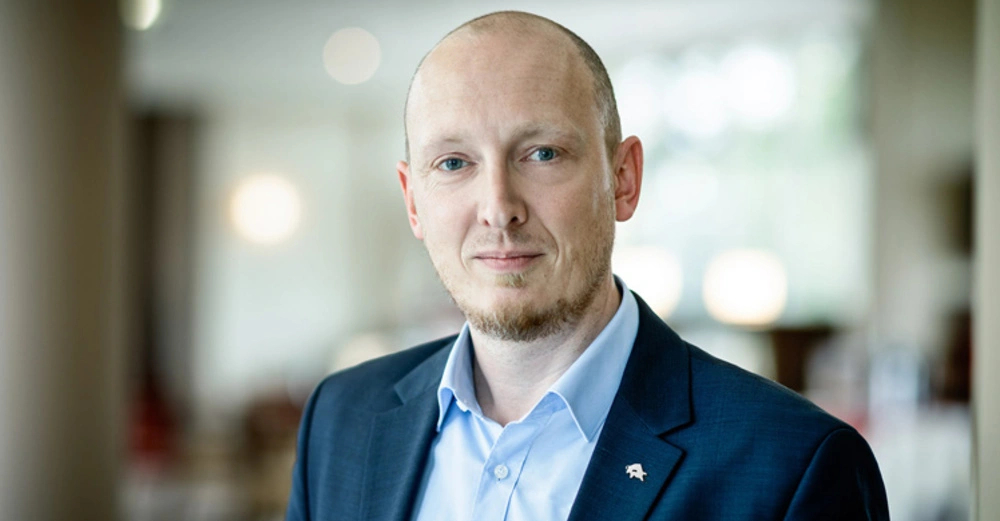 Mann trägt einen Anzug mit hellblauem Hemd steht ruhig in einem hellen Raum mit verschwommenem Hintergrund aus Licht und Möbeln