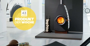 Ein Kaminofen brennt auf einem Podest in einem modernen Wohnzimmer. Daneben steht ein Holzstapelhalter. Links ein großes Kunstwerk. Text: Kalender mit Nummer 43 PRODUKT DER WOCHE.