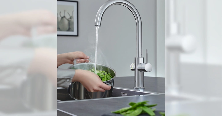 Hände halten einen Topf mit frischem Gemüse unter fließendes Wasser aus einem modernen Wasserhahn in einer Küche mit grauem Interieur und einem Bild an der Wand.