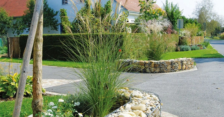 Ein mit Steinen gefüllter Metallkorb steht neben einem Baum in einem begrünten Verkehrsbereich mit Zierpflanzen. Im Hintergrund sind ein Haus mit rotem Dach und eine Straße zu sehen.