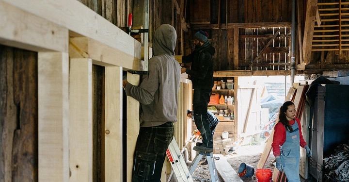 2 Studenten von BASEhabitat stehen auf Leitern und fixieren eine Holzbalkenkonstruktion. Studentin in blauer Latzhose und rotem Pulli geht durch den Raum.