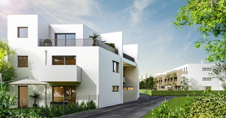 Ein modernes weißes Gebäude steht ruhig an einer asphaltierten Straße umgeben von grünen Bäumen und Pflanzen. Im Hintergrund sind weitere ähnliche Wohnhäuser zu sehen.