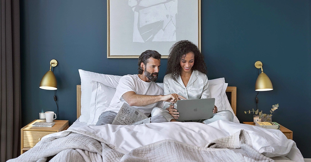 Ein Mann und eine Frau sitzen im Bett und schauen auf einen Laptop Dunkelblaue Wände umgeben das Bett auf Nachttischen links und rechts stehen Lampen Bücher und dekorative Gegenstände