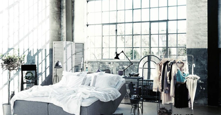 Ein Doppelbett mit weißer Bettwäsche steht vor einer großen Fabrikfensterwand in einem modern eingerichteten Loft mit Pflanzen Lampen und einem Kleiderständer voller Kleidung.