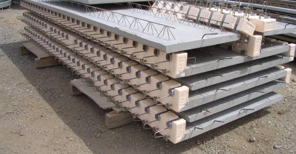 Mehrere gestapelte Betonplatten mit Drahtbündeln auf einer Baustelle auf hölzernen Stützen gelagert in einem industriellen Umfeld im Freien.