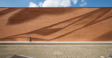 Person geht vor großem rotem Backsteingebäude mit wellenförmigen Mustern spazieren Kontext klarer Himmel mit wenigen Wolken und gepflasterter Boden.