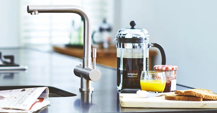Ein Wasserhahn aus Edelstahl steht still auf einer Küchenarbeitsplatte neben einer Zeitung, einer French Press, einem Glas Orangensaft und einem Schneidebrett mit Marmelade und Toast.