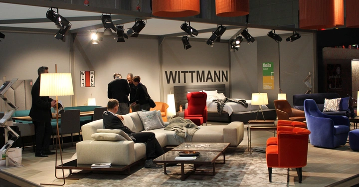 Galerie-Bild 12: Mehrere Möbel wie Sofas und Sessel sind in einem Ausstellungsraum arrangiert mit Menschen, die die Möbel betrachten. Der Hintergrund zeigt den Markennamen WITTMANN und Lampen hängen von der Decke.