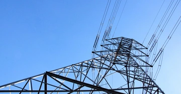 Strommast trägt Hochspannungsleitungen aufwärts in Richtung klaren blauen Himmel. Kabel erstrecken sich vom Turm in verschiedene Richtungen. Umgebung wirkt frei von Hindernissen und unbebaut.