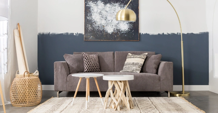 Galerie-Bild 6: Graues Sofa steht vor einer zweifarbigen Wand unter einem Kunstwerk; daneben eine goldene Bogenlampe; zentral zwei runde Tische auf einem Teppich; links ein Korb und gerollte Poster.