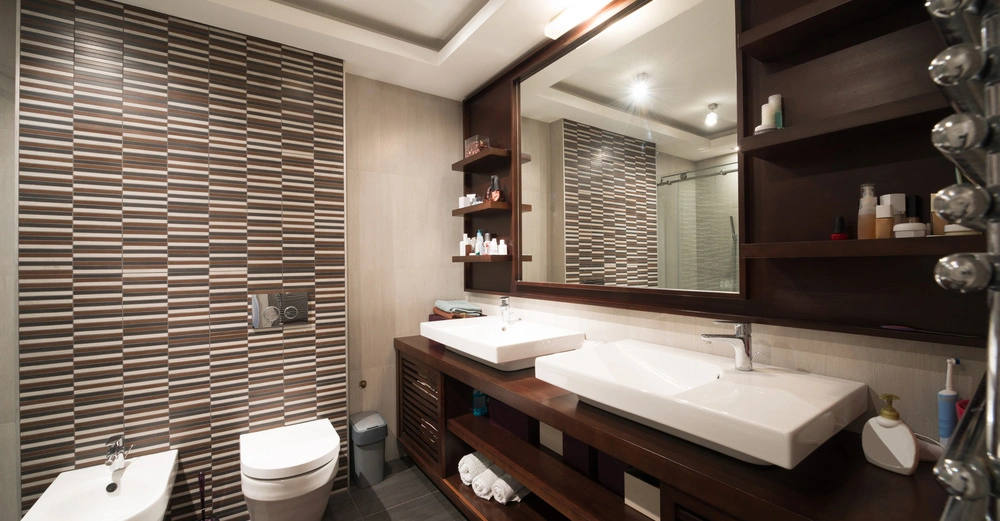 Zwei Waschbecken auf einem Holztisch mit einem großen Spiegel darüber befinden sich in einem modernen Badezimmer mit vielen Regalböden auf der rechten Seite und einer Toilette sowie einem Bidet links.