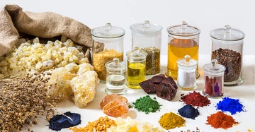 Verschiedene Gewürze und farbige Pulver liegen auf einem weißen Tisch vor mehreren Glasgefäßen mit Deckeln. Ein Sack mit gelben Kristallen ist auf der linken Seite zu sehen.