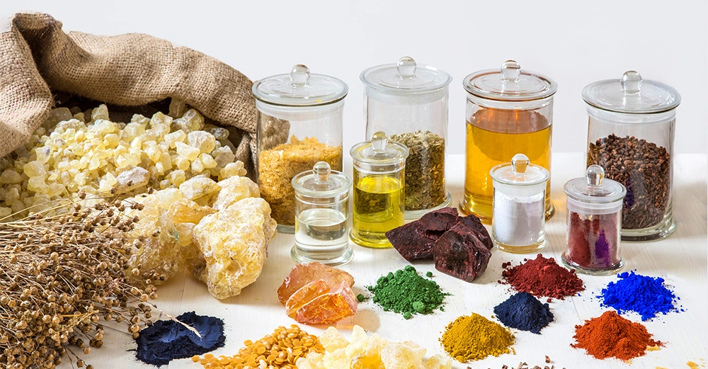 Verschiedene Gewürze und farbige Pulver liegen auf einem weißen Tisch vor mehreren Glasgefäßen mit Deckeln. Ein Sack mit gelben Kristallen ist auf der linken Seite zu sehen.