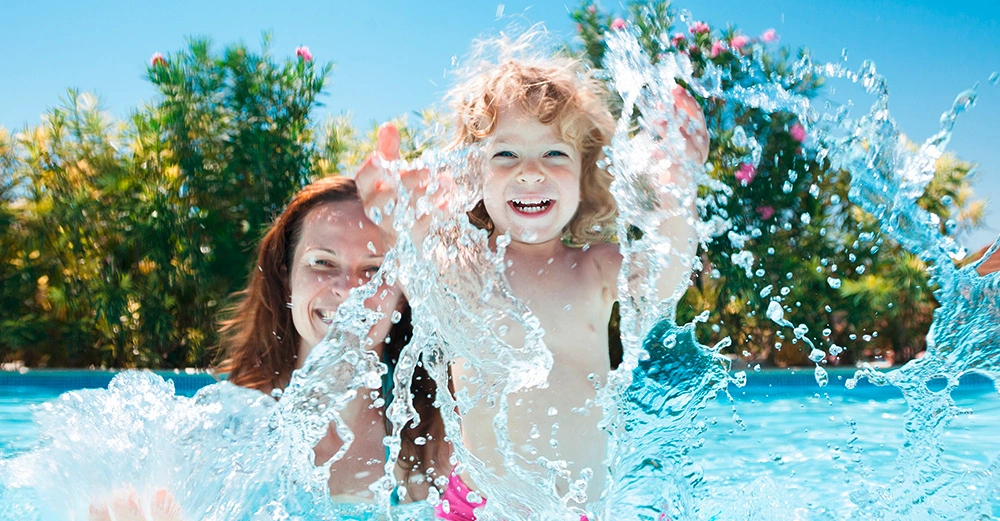 Kind und Erwachsener spielen und lachen im Pool umgeben von blühenden Büschen und klarem Himmel Hintergrund.