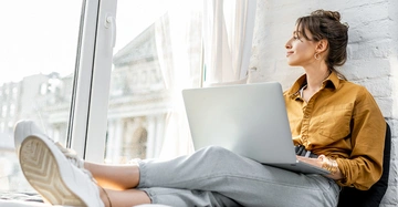 Eine Frau benutzt einen Laptop und sitzt entspannt an einem großen Fenster. Sie trägt ein gelbbraunes Hemd und graue Hosen. Draußen sind verschwommene Gebäude zu sehen.