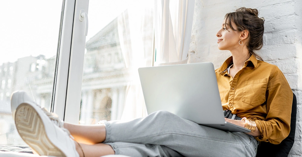 Eine Frau benutzt einen Laptop und sitzt entspannt an einem großen Fenster. Sie trägt ein gelbbraunes Hemd und graue Hosen. Draußen sind verschwommene Gebäude zu sehen.