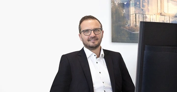 Mann sitzt lächelnd am Schreibtisch trägt Brille und Anzug im Hintergrund Bild eines modernen Gebäudes Monitor im Vordergrund büroumgebung