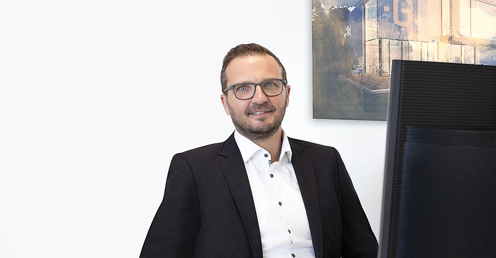 Mann sitzt lächelnd am Schreibtisch trägt Brille und Anzug im Hintergrund Bild eines modernen Gebäudes Monitor im Vordergrund büroumgebung