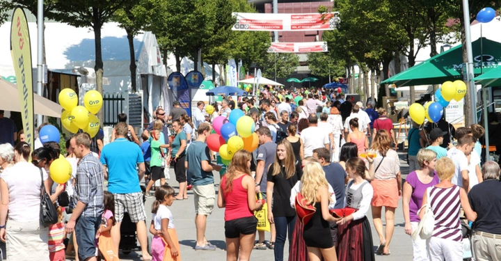 Menschenmenge spaziert auf einer breiten Straße bei einer Veranstaltung. Viele tragen bunte Luftballons mit der Aufschrift ÖVP. Umgeben von Informationsständen und grünen Bäumen. Banner im Hintergrund.