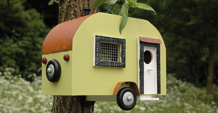 Galerie-Bild 2: Ein kleines Vogelhaus in Form eines Wohnwagens hängt an einem Baum in einem bewaldeten Garten. Der Wohnwagen hat ein Fenster und eine Tür, die als Eingang für Vögel dient.