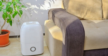 Ein weißer Luftentfeuchter steht auf einem Boden neben einem braunen Sofa und einer Grünpflanze im roten Blumentopf vor einer weißen Wand.