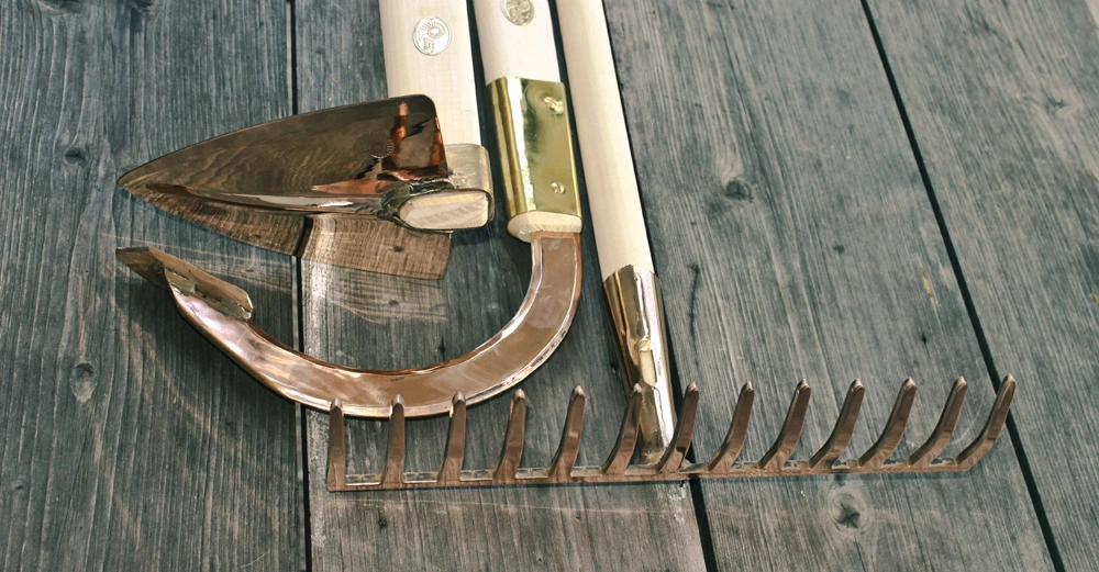 Metallene Gartenwerkzeuge liegen auf einem verwitterten Holztisch. Ein Spaten eine Laubharke und ein Sichelmesser sind dicht nebeneinander gruppiert. Es gibt kein Text im Bild.
