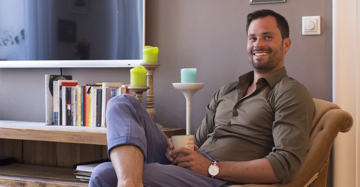 Ein Mann sitzt lächelnd auf einem Sessel und hält eine Tasse. Er befindet sich in einem Wohnzimmer mit Büchern, einer großen abgeschalteten TV und mehreren gerahmten Bildern an der Wand.