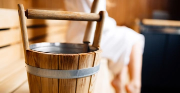 Holzeimer steht still in einer Sauna Umgebung mit Holzbänken wo eine Person in einem weißen Bademantel sitzt.