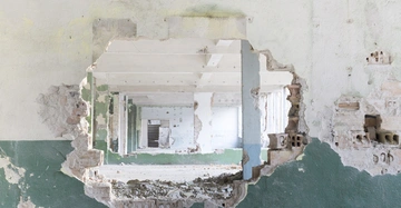 Ausgebrochene Mauer mit vielen Löchern führt zu leeren, heruntergekommenen Räumen hintereinander, zerbrochene Ziegelsteine und Schutt auf dem Boden, grün-weiße Wandfarbe teilweise abgeblättert in einem verlassenen Gebäude.