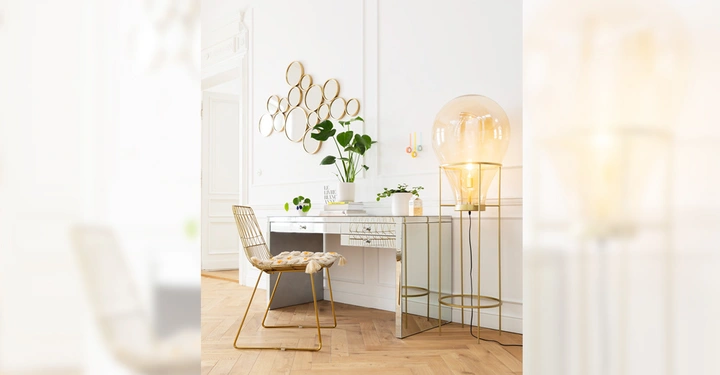 Galerie-Bild 3: Ein goldener Stuhl steht vor einem weißen Schreibtisch in einem hellen Raum zwei Pflanzen und eine große Lampe auf dem Boden zieren die Umgebung an der Wand hängen runde Spiegel.