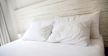 Bett mit weißer Bettwäsche und Kissen steht in einem Schlafzimmer mit weiß gestrichener Holzwand im Hintergrund bei Tageslicht.