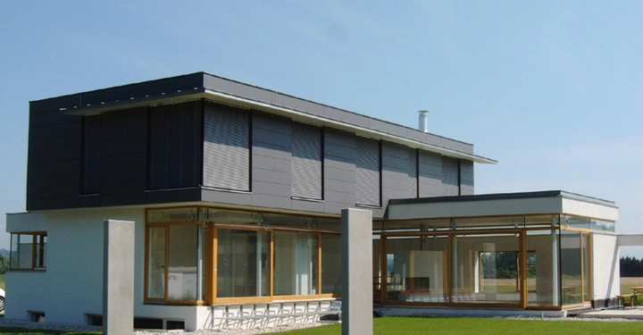 Modernes zweistöckiges Haus mit großen Fenstern und grauen Jalousien steht auf einem gepflegten grünen Rasen an einem sonnigen Tag.