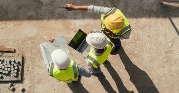 Arbeiter in Bauhelmen und Warnwesten begutachten Pläne und einen Laptop auf einer Baustelle im Freien wobei einer auf etwas in der Ferne zeigt.