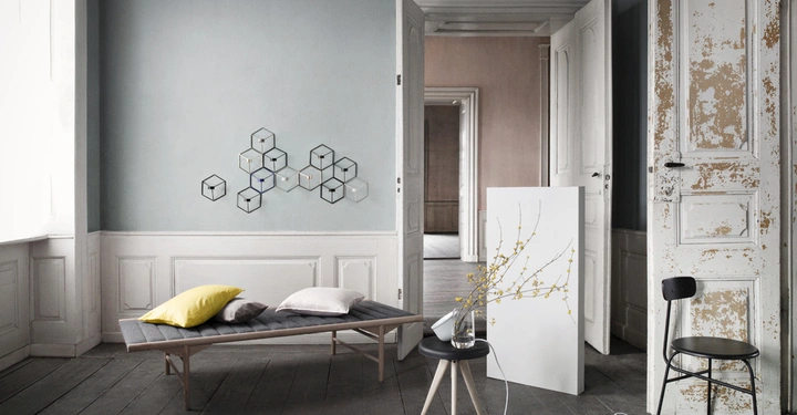 Galerie-Bild 11: Ein graues Sofa mit Kissen steht in einem Raum mit Holzfußboden und grauen Wänden. An der Wand befinden sich geometrische Wanddekorationen, ein Stuhl und ein kleines Tischchen mit Vase und Zweig.