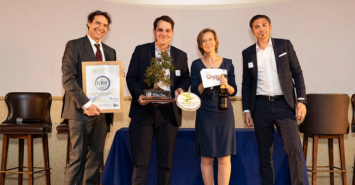Galerie-Bild 2: Vier Personen stehen nebeneinander und halten Auszeichnungen und Preise, darunter ein gerahmtes Zertifikat, eine kleine Pflanze und eine Flasche Sekt, in einem formellen Raum. Text: URKUNDE GBB 2021 Nature One Ghezzo GBB AWARDS 2021