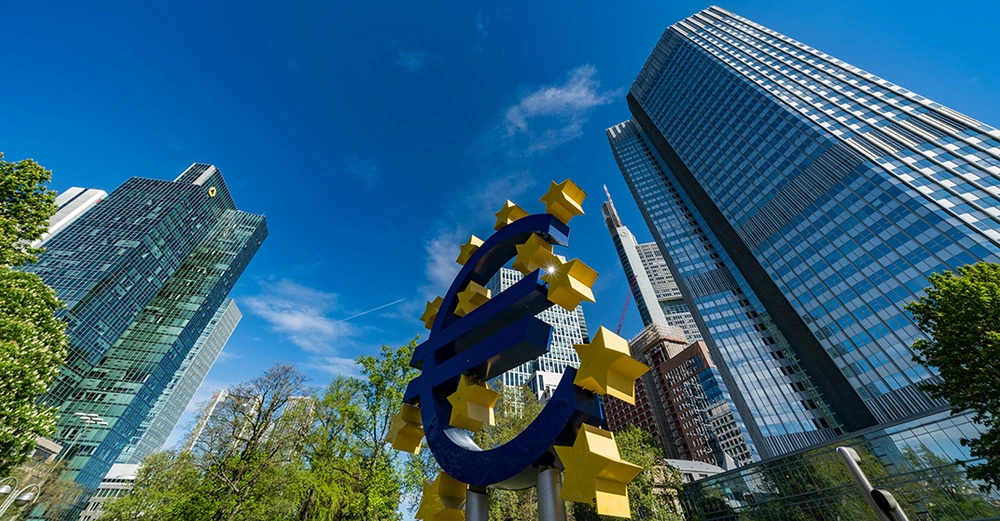 Große Euro-Skulptur aus blauen Röhren und gelben Sternen stehend in einem modernen Stadtgebiet umgeben von hohen Bürogebäuden und Bäumen unter einem klaren blauen Himmel.