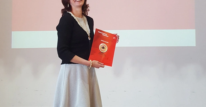 Eine Frau lächelt und hält eine rote Mappe auf einem Holzfußboden in einem Raum. Der Hintergrund zeigt eine Präsentationsfolie mit dem Text 37 ordentliche Generalversammlung Österreichischer Fertighausverband