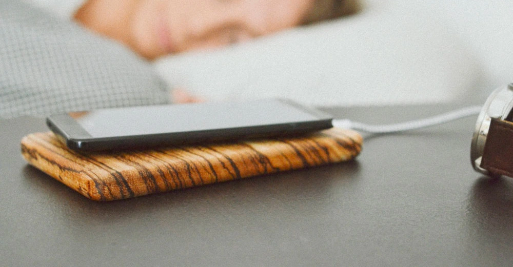 Smartphone ruht auf Holzladegerät am Nachttisch daneben Armbanduhr im Hintergrund schlafende Person im Bett