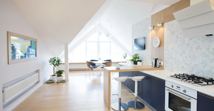 Ein moderner Küchenbereich mit Herd und zwei Barhockern befindet sich in einem hellen Raum, der zu einem Essbereich mit einem runden Tisch und vier Stühlen unter einer großen Dachgaube führt.