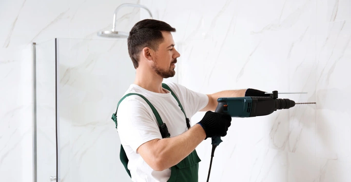 Mann in weißem T-Shirt und grüner Latzhose bohrt mit einer elektrischen Bohrmaschine ein Loch in eine geflieste Wand in einem Badezimmer mit Duschkabine.