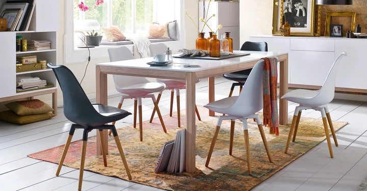 Galerie-Bild 1: Holztisch umgeben von bunten Stühlen steht auf einem Teppich mehrere Dekorationsgegenstände und Bücher auf dem Tisch und in Regalen große Fenster im Hintergrund lassen Licht hinein helle und moderne Wohnungseinrichtung