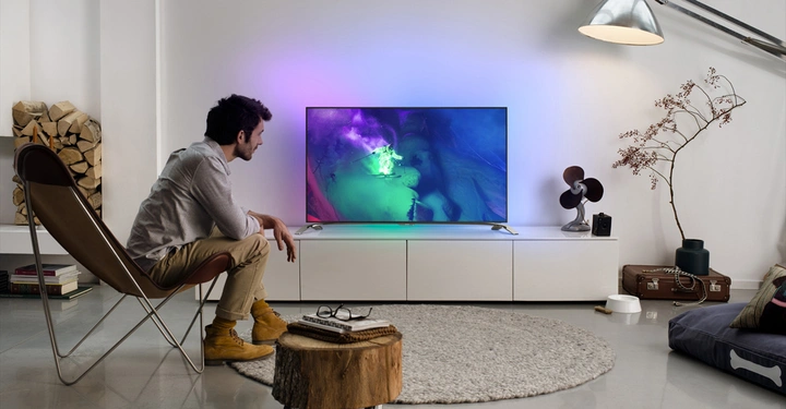 Galerie-Bild 1: Ein Mann sitzt auf einem Stuhl und schaut auf einen eingeschalteten Fernseher in einem modern eingerichteten Wohnzimmer mit Holzscheiten, einem Teppich und einer Stehlampe.