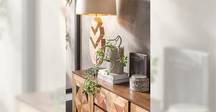 Eine Lampe leuchtet auf einem Holztisch stehend neben einem Buch mit der Aufschrift THE KINFOLK ENTREPRENEUR und einer Topfpflanze die vom Tisch herunterhängt in einem modern eingerichteten Raum