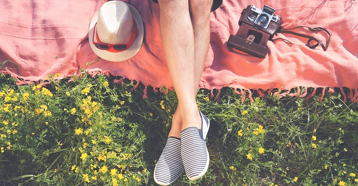 Beine liegen auf einer Decke im Gras mit gelben Blumen Hut und Sonnenbrille links und alte Kamera rechts in heller Umgebung